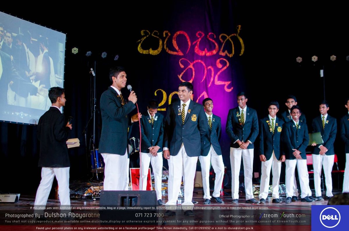 Association members in school blazers on stage beside a presenter with a microphone, in front of a Sahasak Naada backdrop