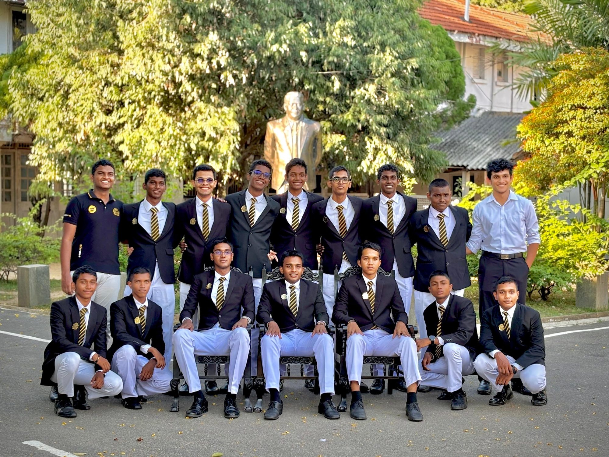 The top board of The Radio Electronics Association of D. S. Senanayake College in school blazers, standing and seated in front of a statue in the school grounds